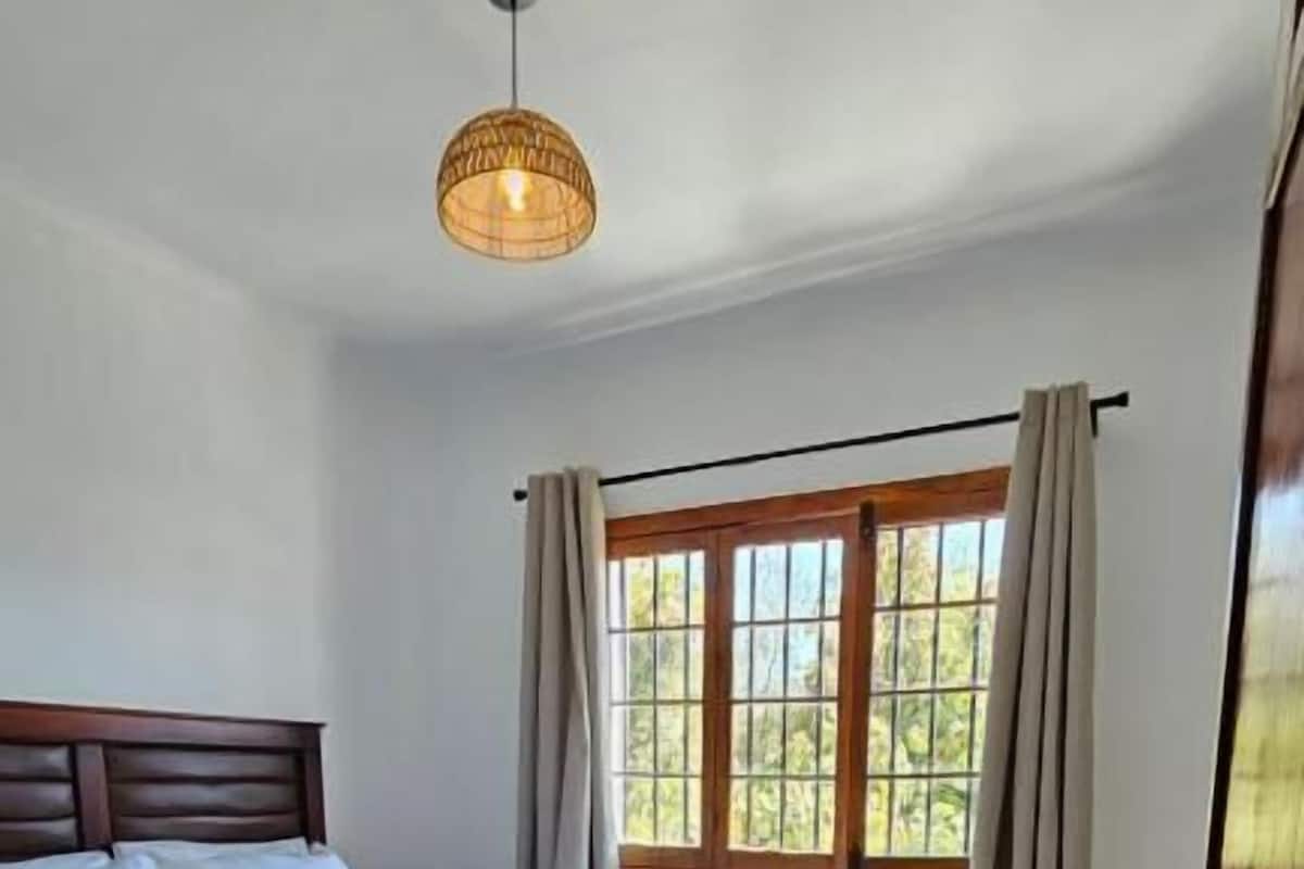A bedroom with a wooden bed, a hanging light fixture, a window with curtains, and a door with a key.