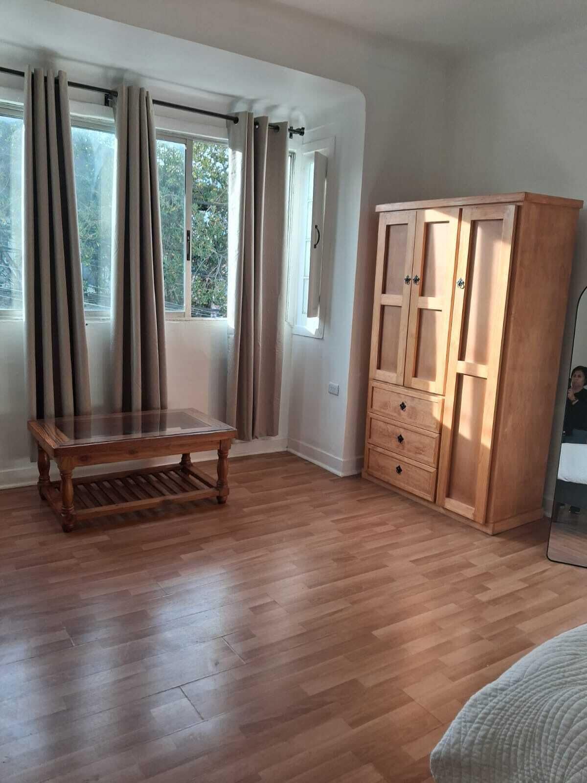 A room with wooden flooring, a wooden table, a wardrobe, and a window with curtains.