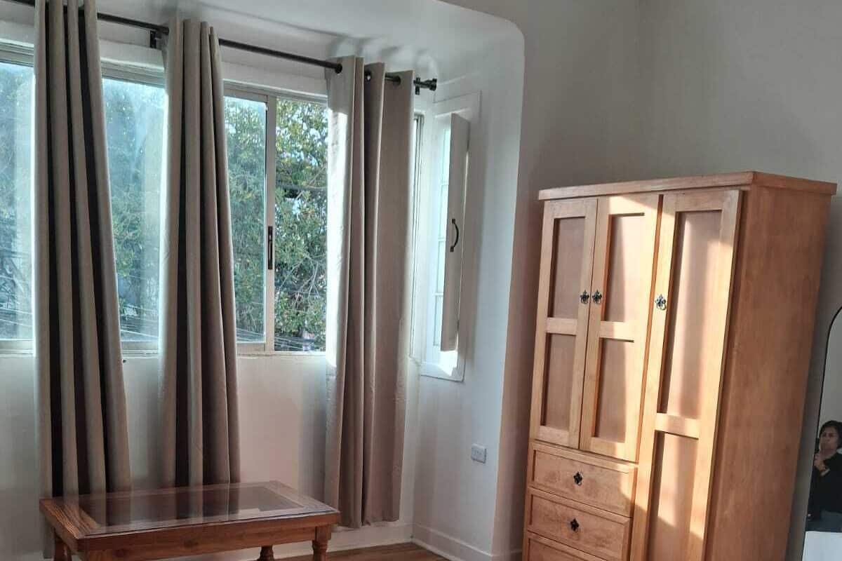 A room with wooden flooring, a wooden table, a wardrobe, and a window with curtains.