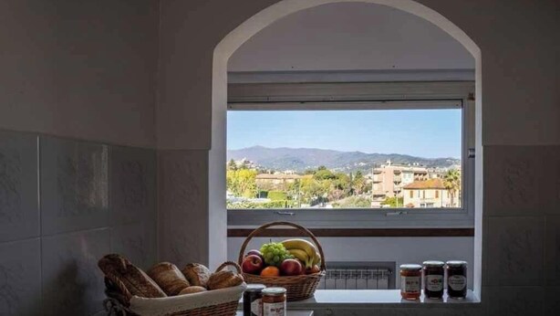 Una cocina con vistas a través de una ventana arqueada, una cesta de pan, frutas y botes de conservas.