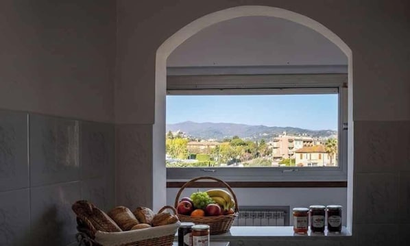 A kitchen with a view through an arched window, a basket of bread, fruits, and jars of preserves. - Holiday apartment with air conditioning and balcony with sea view (Diano Marina)