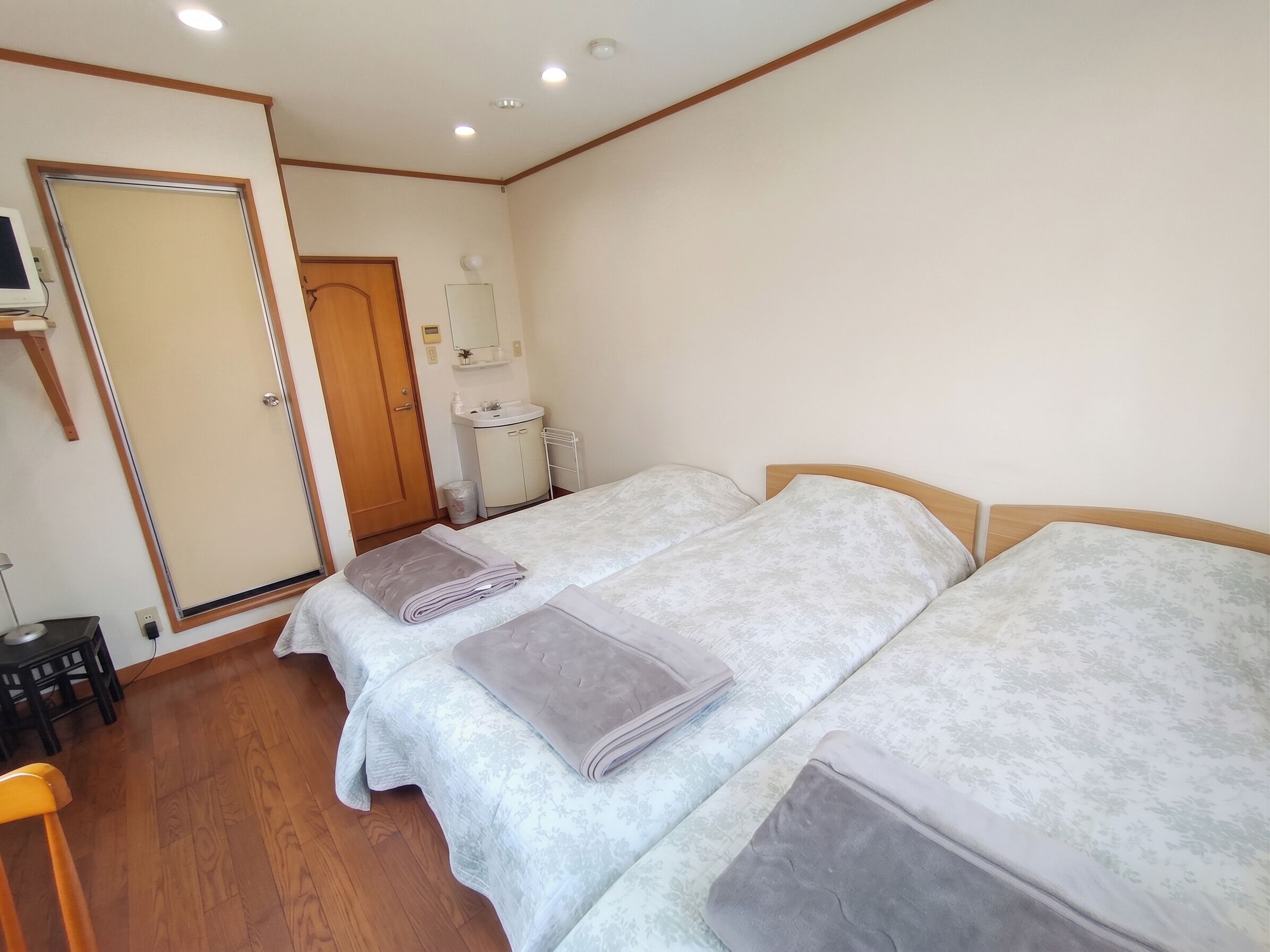 Habitación de hotel con dos camas, suelo de madera, baño con lavamanos y espejo, y una puerta que da a otra habitación.