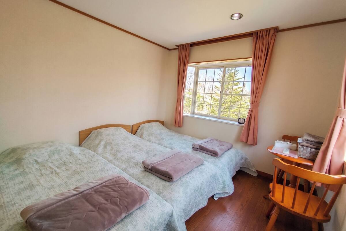 A bedroom with two beds, a wooden chair, a window with curtains, and a small table with items on it.