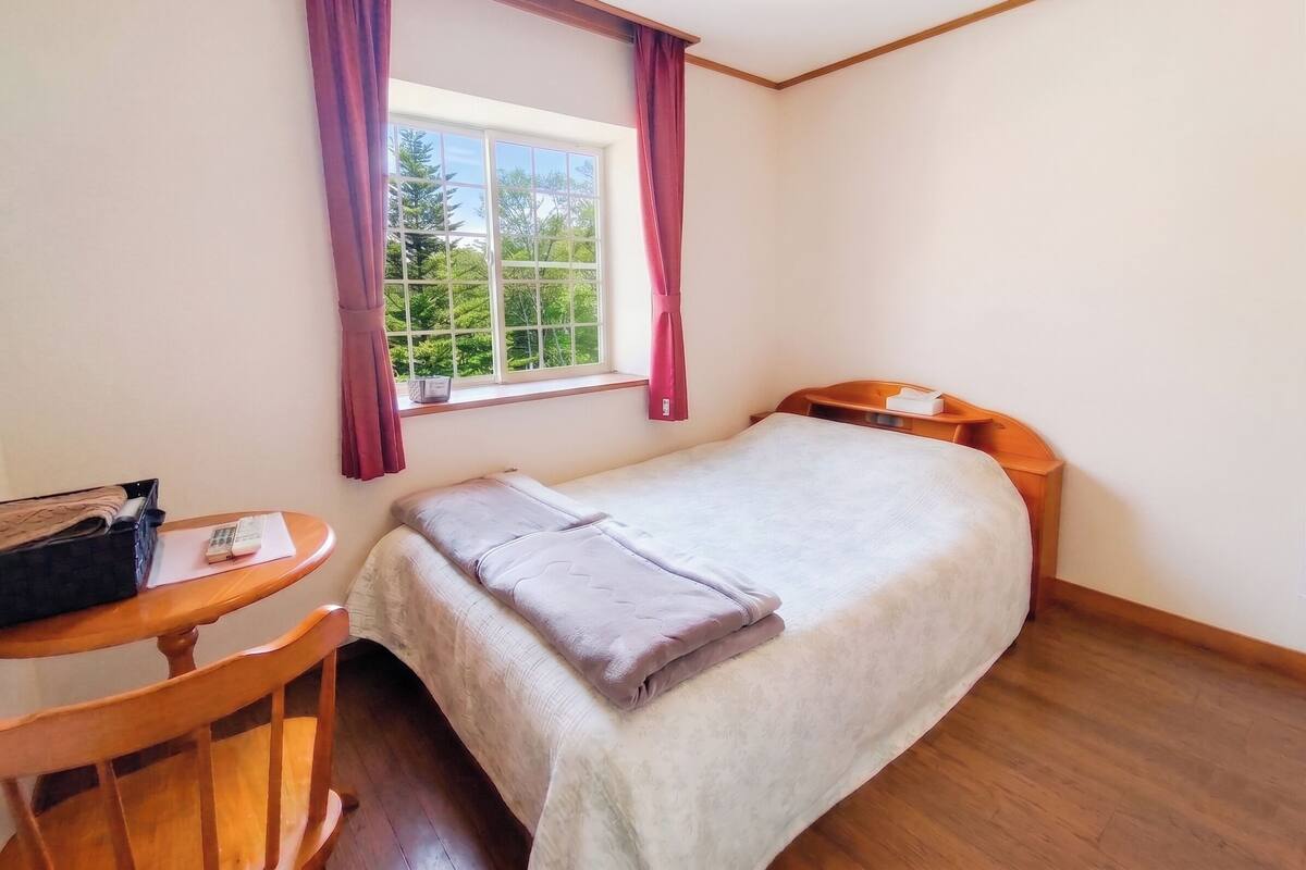 A room with a bed, a small table, a window with curtains, and a view of greenery outside.