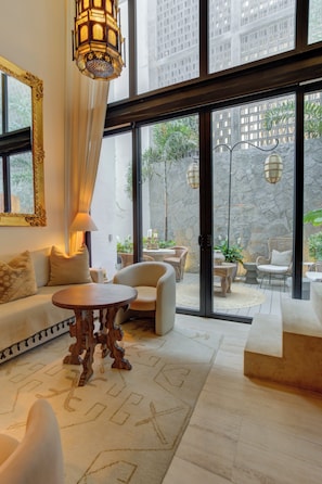 Ein geräumiges Wohnzimmer mit Sofa, Sesseln, einem Couchtisch und einem großen Fenster mit Blick auf einen Innenhof mit Pflanzen und einer Steinmauer.