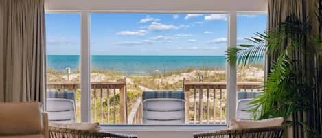 A room with a large window overlooking a beach, wicker chairs, a small round table, and a potted plant.