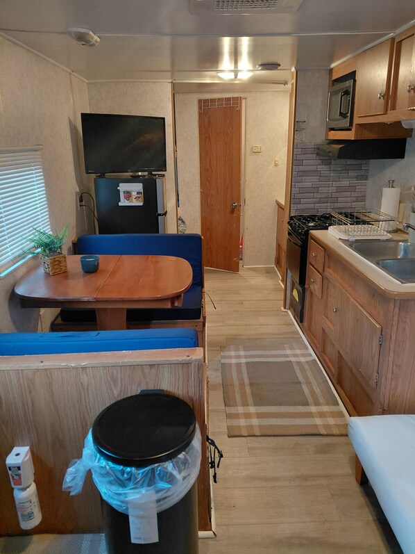A compact RV kitchen with a dining area, a trash can, and a small refrigerator.