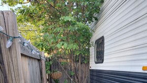 Une clôture en bois, une petite terrasse avec une table et des chaises, une plante suspendue et un camping-car blanc.