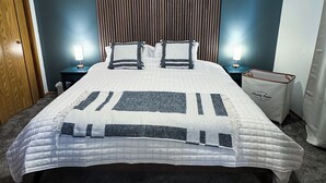 A neatly made bed with a quilted bedspread, two bedside tables with lamps, a wooden wardrobe, and a laundry basket.