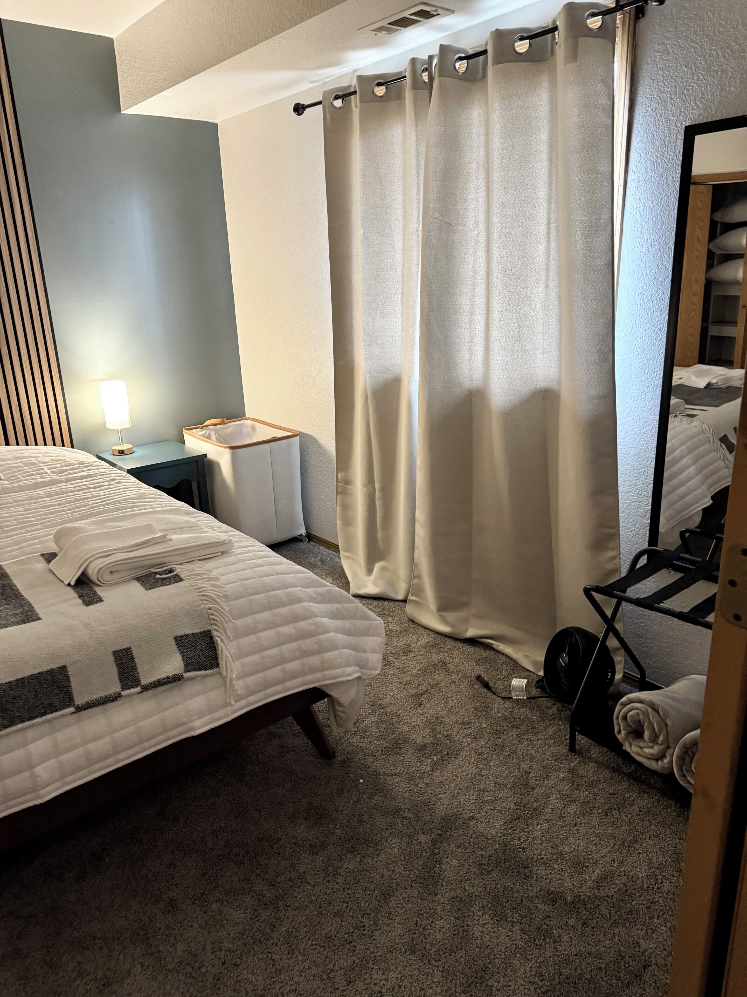 A hotel room with a bed, a bedside table with a lamp, a small table, a fan, and a mirror.