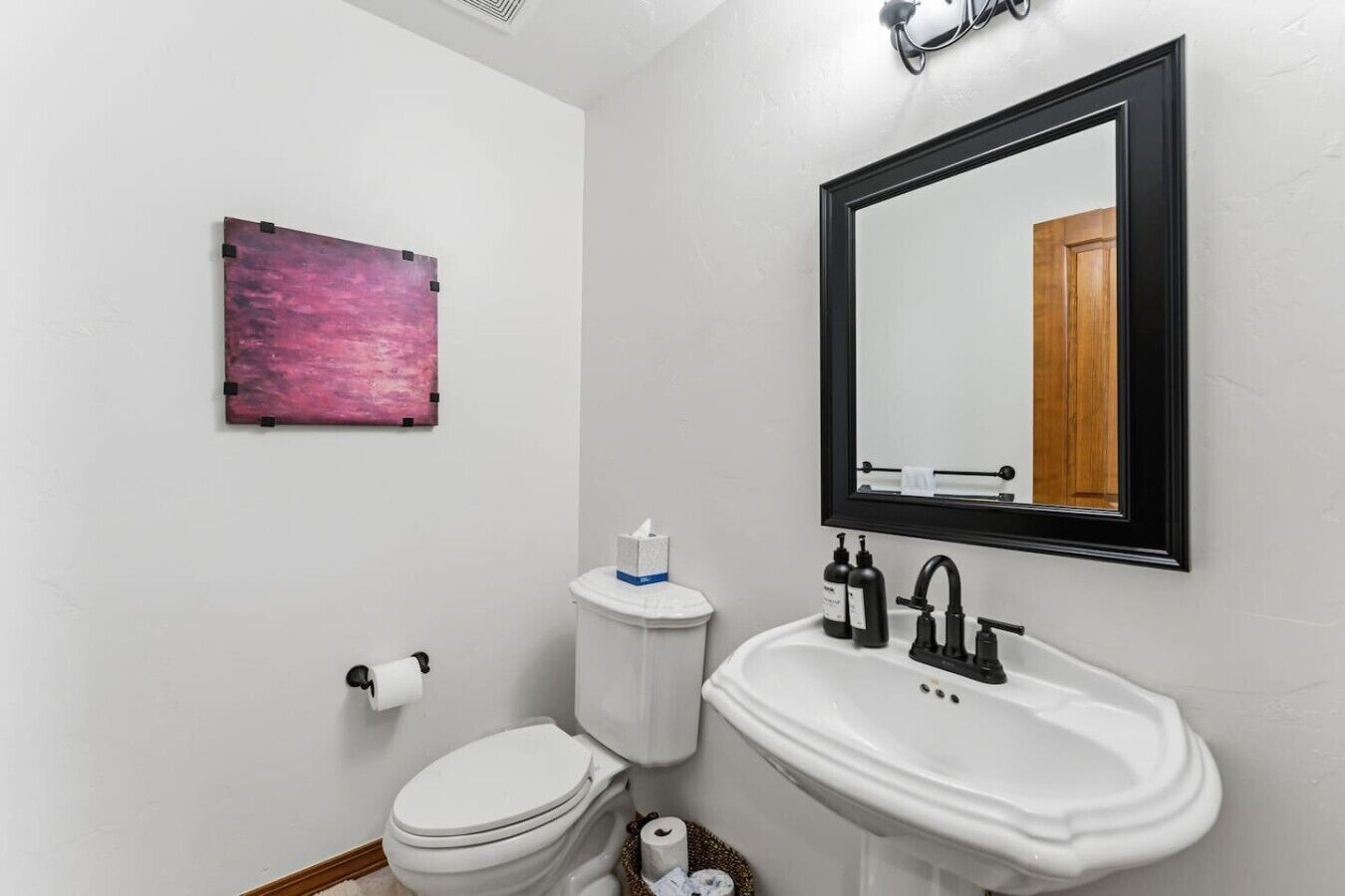 Un baño con inodoro, lavamanos, espejo y un cuadro abstracto rosa en la pared.