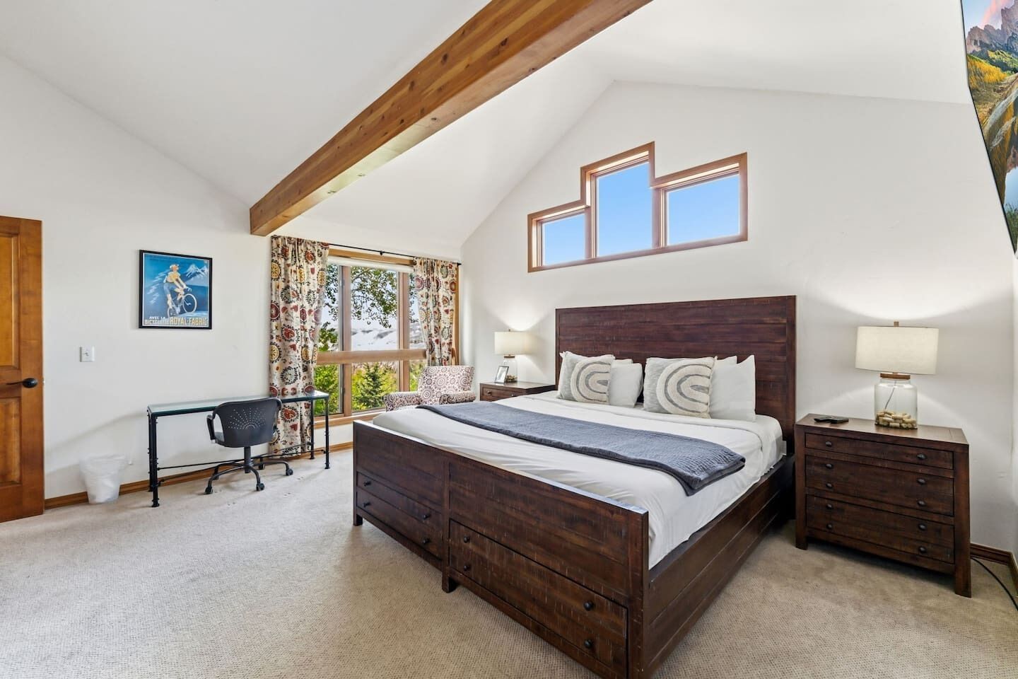 A bedroom with a wooden bed, a desk, and a chair. There are three windows and a door.