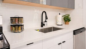A modern kitchen with a stainless steel sink, a black faucet, and a dish soap dispenser. There are spice jars on the counter and a potted plant. - Modern Miami Retreat w/ Games (Miami)