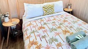 A hotel room with a bed, two bedside lamps, a nightstand with a vase of flowers, and a wall-mounted air conditioner. - Summer – vintage retro caravan, sleeps 5 | Rye, Mornington Peninsula (Rye)