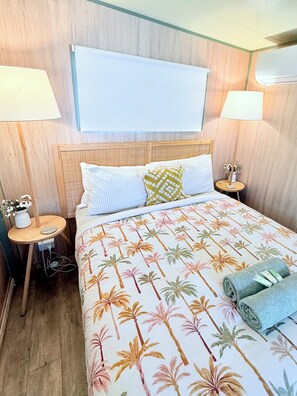 Ein Hotelzimmer mit einem Bett, zwei Nachttischlampen, einem Nachttisch mit Blumenvase und einer Wand-Klimaanlage.