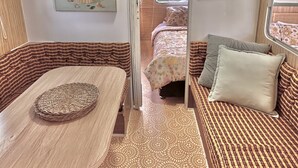 A compact RV interior with a dining area, a bed with patterned bedding, and a small table with a woven placemat. - Summer – vintage retro caravan, sleeps 5 | Rye, Mornington Peninsula (Rye)