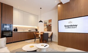A modern kitchen with a dining area, a large screen displaying 'Grandstay Collection,' and a cozy seating arrangement. - Grandstay Sea Penthouse 04 (Durrës)