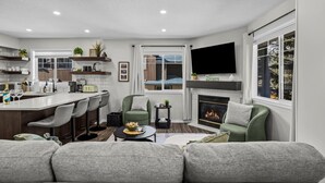 A modern living room with a sofa, a dining area with bar stools, a fireplace, and a TV mounted on the wall. - Cozy Rockies Stay with Hot Tub and Fire Pit (Harvie Heights)