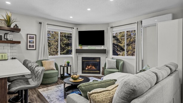 A modern living room with a fireplace, green and grey furniture, a flat-screen TV, and a dining area with a white table and bar stools. - Cozy Rockies Stay with Hot Tub and Fire Pit (Harvie Heights)