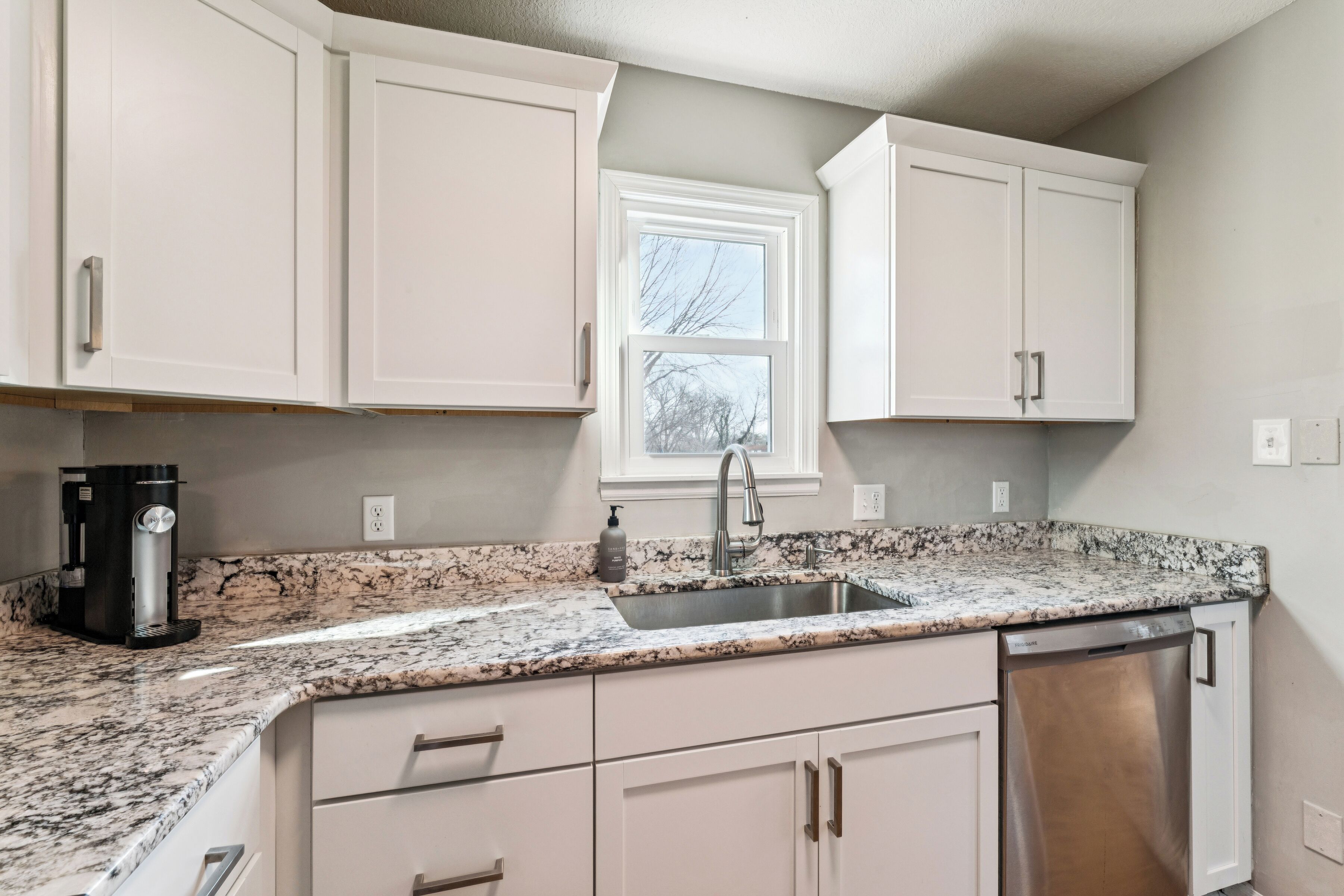 A kitchen with white cabinets, a granite countertop, a stainless steel sink, a coffee maker, and a dishwasher.