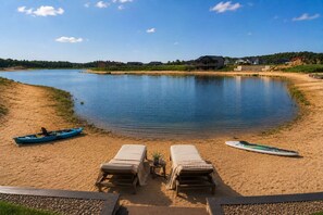 A lakeside view with two lounge chairs, a kayak, and a paddleboard on the sand. - Jimmy’s Beach House | Private Beach + Arcade (New Lisbon)