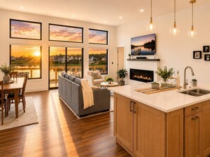 A modern kitchen with a fireplace, wooden cabinets, and a dining area with a view of a sunset. - Jimmy’s Beach House | Private Beach + Arcade (New Lisbon)
