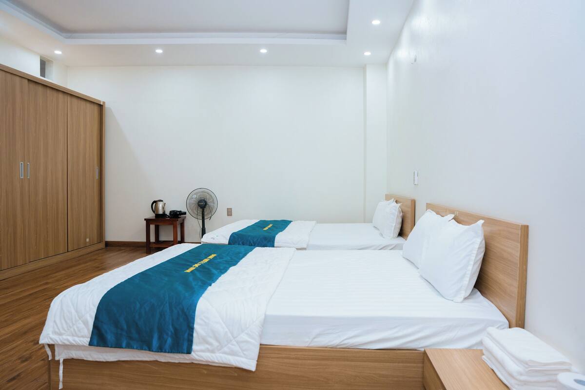 Habitación de hotel con dos camas, un armario de madera, una mesita con una tetera y un ventilador.