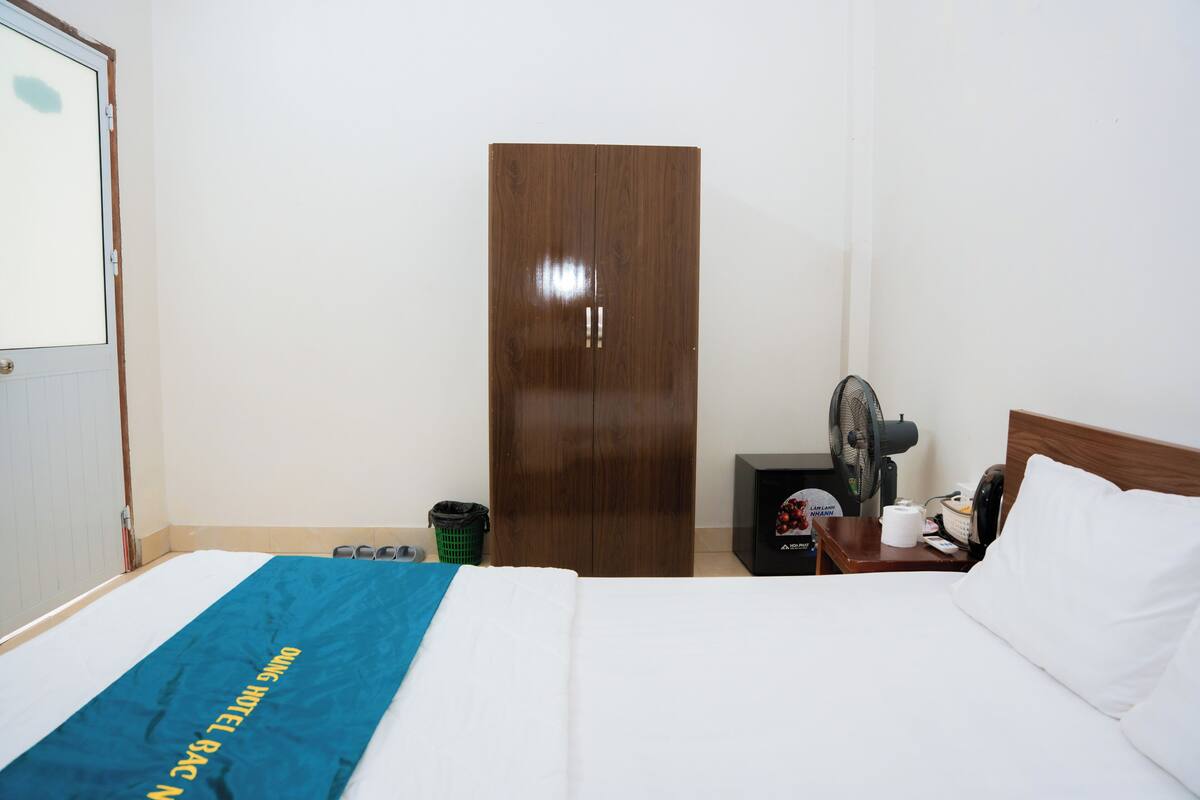 Una habitación de hotel con una cama, un armario de madera, una mesita de noche con un ventilador y un cubo de basura.