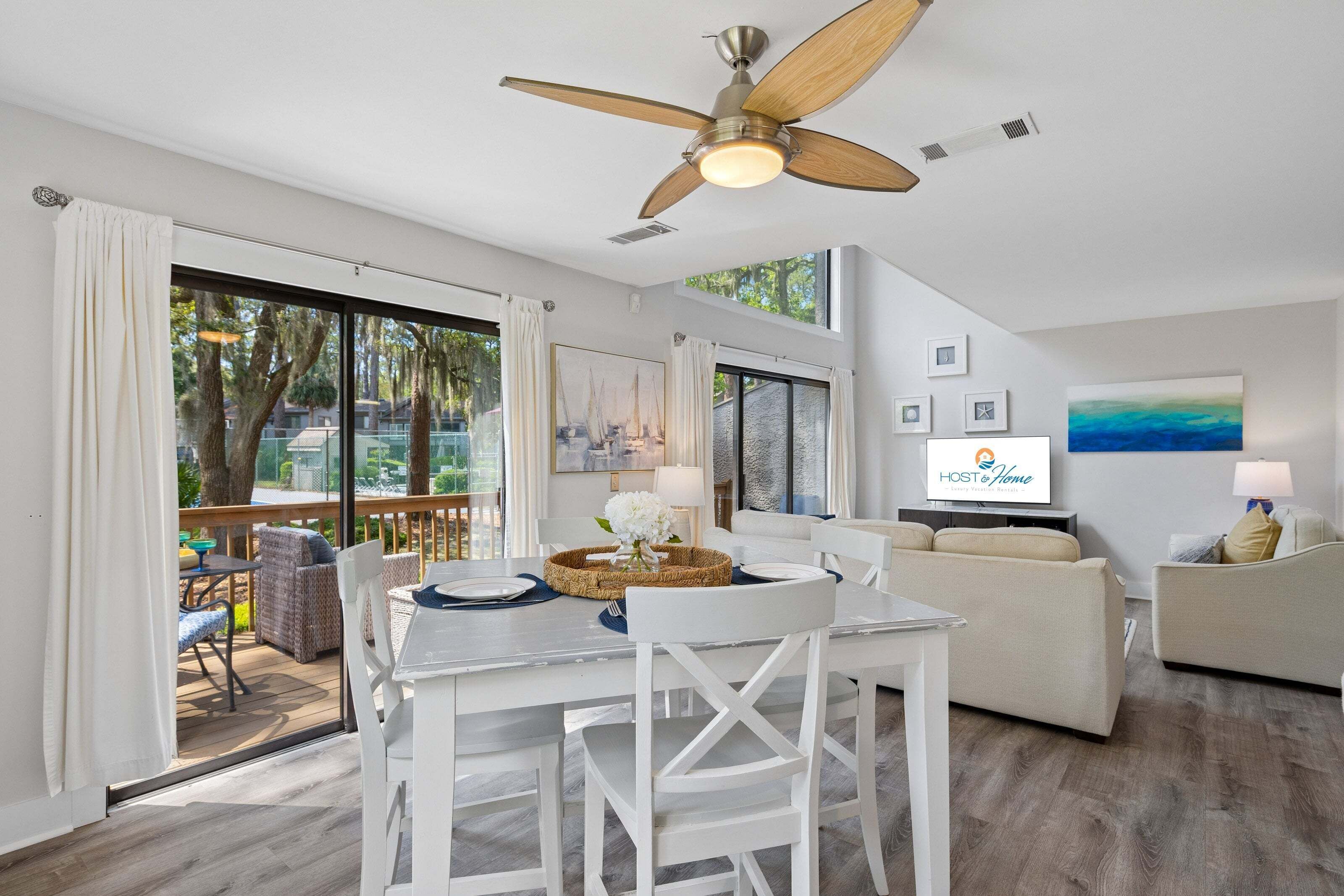 A modern living room with a dining area, a ceiling fan, large windows, and a TV.