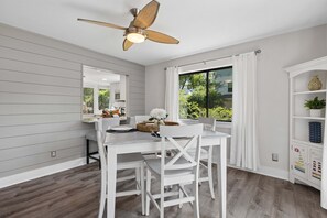 A dining area with a white table and chairs, a ceiling fan, a large window with curtains, and a cabinet with decorative items. - Forest Beach | Walk to Beach, Pool & Tennis, Private Balcony, Sleeps 6 (Hilton Head Island)