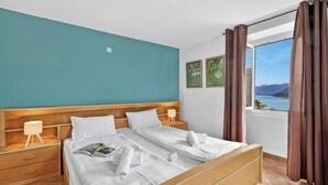 A hotel room with a large bed, two bedside tables, a window with curtains, and a view of the sea. - Casa Berta Brissago 1 with Balcony for Four, Brissago, Switzerland (Brissago)