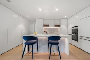 A modern kitchen with white cabinetry, a marble countertop, two blue bar stools, and built-in appliances. - 海滨CBD/奢华2室/可住6人/海港大桥/悉尼歌剧院 (Barangaroo)