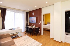 A modern hotel room with a bed, a sofa, a dining area with a table and chairs, a flat-screen TV mounted on the wall, and a view of the cityscape through the window. - La Dimora Azzurra 102, Vlore, Albania (Vlore)