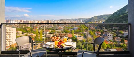 A balcony with a table set for breakfast, overlooking a cityscape with buildings and hills.
