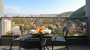 Un balcón con mesa lista para el desayuno, con vistas a un paisaje urbano de edificios y colinas.