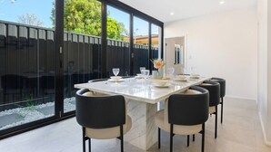 A modern dining area with a large table, black chairs, and a view of a backyard through large windows. - Spacious 5BR House for 14ppl/Family Gathering/Park (Kingsgrove)