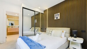 A modern hotel room with a large bed, a bedside table with a lamp, a mirror, and a wooden panel wall. - Spacious 5BR House for 14ppl/Family Gathering/Park (Kingsgrove)
