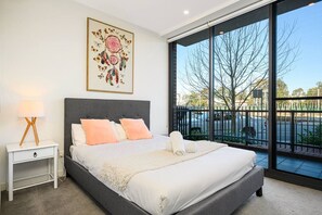 A bedroom with a large bed, a bedside table with a lamp, a framed artwork, and a view of the outside.