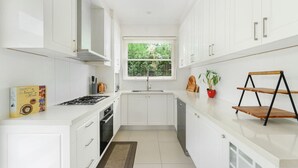 Une cuisine étroite avec des meubles blancs, une fenêtre avec des stores, une cuisinière à gaz et une petite étagère sur laquelle est posée une plante.