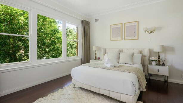 Une chambre avec une grande fenêtre, un lit, deux tables de chevet et des œuvres d’art encadrées accrochées au mur.