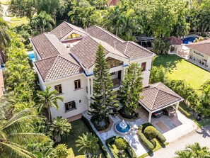 A large, multi-story house with a tiled roof, surrounded by lush greenery and a swimming pool.