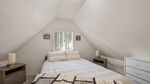 Un dormitorio con techo inclinado, una cama con ropa de cama blanca, dos mesitas de noche con lámparas, un aparador y una ventana con persianas.