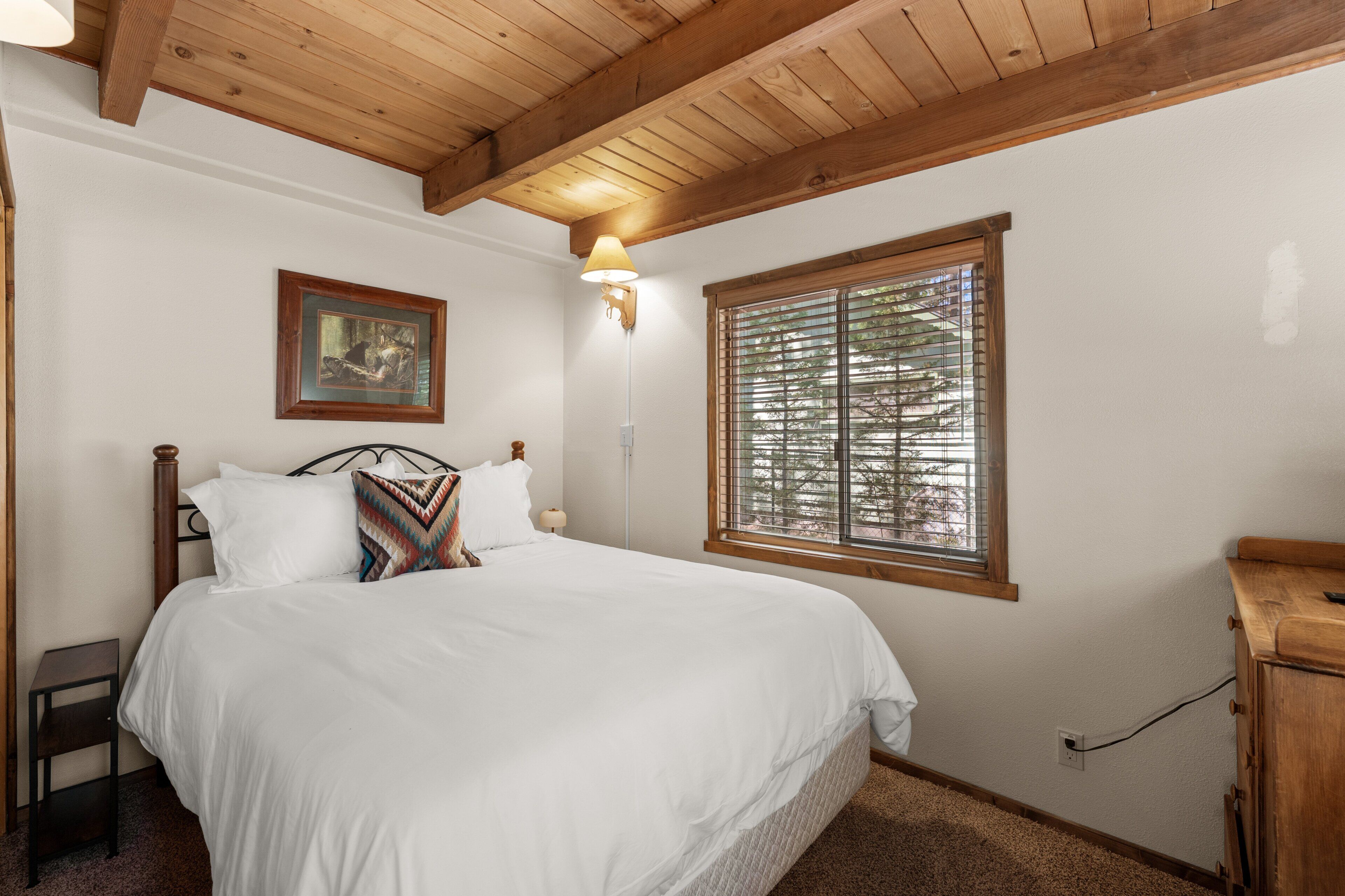 Une chambre avec un plafond en bois, un lit avec une literie blanche, une table de chevet, une lampe fixée au mur, une fenêtre avec des stores et un tableau encadré accroché au mur.