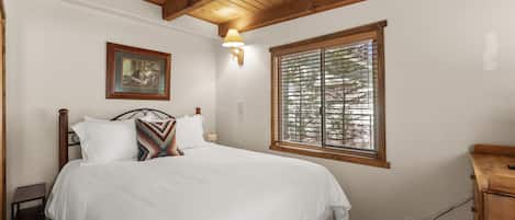 Quarto com teto de madeira, cama com roupa de cama branca, mesa de cabeceira, luminária na parede, janela com persianas e um quadro na parede.