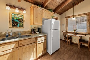 A kitchen with wooden cabinets, a white refrigerator, granite countertops, a sink, and a dining area with a table and chairs. - Bear Hug Hideaway - 16-Minute walk to Fireworks/Meadow Park (Big Bear Lake)