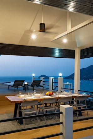 An outdoor dining area with a view of the sea, tables set with plates and glasses, and chairs.