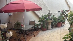 A patio with a table and chairs, a red umbrella, potted plants, and a fountain.