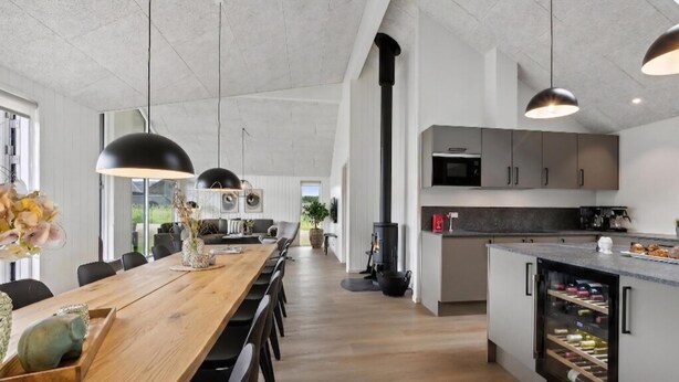 A modern kitchen with a built-in microwave, a wine fridge, and a dining area with a long wooden table and black chairs. - VillaVilla 960 - Østersøbadet, Lolland/Falster/Møn (Rødby)