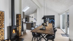 A modern kitchen with a central island, a dining area with a long wooden table and chairs, and a fireplace with a stone facade. - VillaVilla 960 - Østersøbadet, Lolland/Falster/Møn (Rødby)