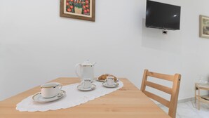 Uma mesa de jantar com um bule, chávenas, pires e um croissant, uma cadeira de madeira, uma televisão na parede e um quadro emoldurado.